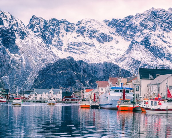 Fiskebåter ligger til havn i Henningsvær. Snøkledde fjell i bakgrunnen.